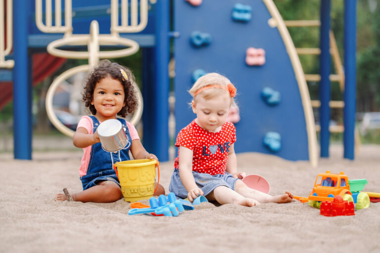 two toddlers sitting in sandbox playing with plastic colorful toys